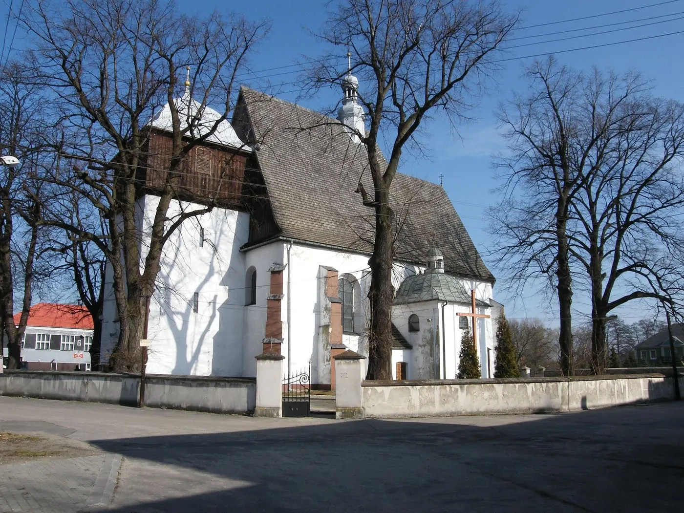 Biały, gotycki kościół z gontowym dachem i drewnianą górną częścią wieży, z kamienno-ceglaną skarpą i białym murem