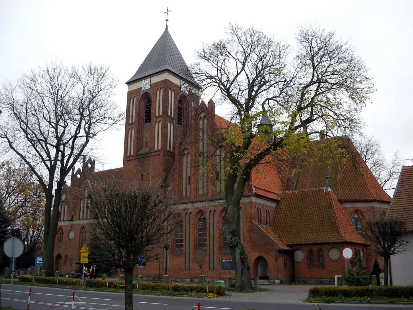 Ceglany, neogotycki kościół z wieżą zwieńczoną szarym, piramidalnym hełmem, widziany od strony ulicy zza bezlistnych drzew; przy drodze znaki i przystanek