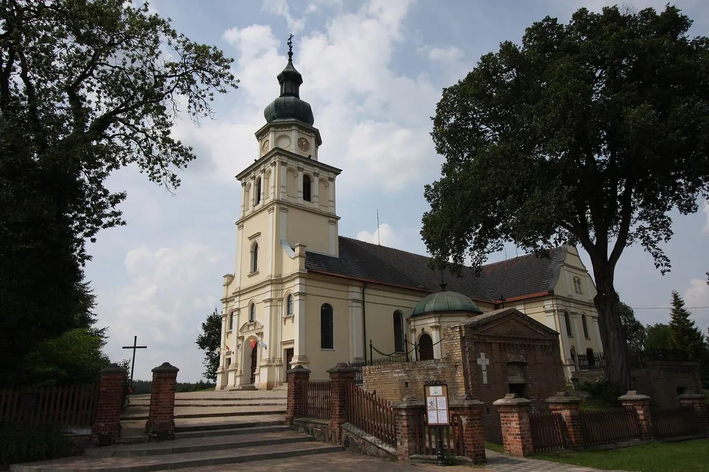 Jasnożółty kościół św. Marii Magdaleny w Pszczewie z wieżą zwieńczoną zielonym, cebulastym hełmem z latarnią i kaplicą nakrytą kopułą, za ceglanymi słupkami bramy, między wysokimi drzewami