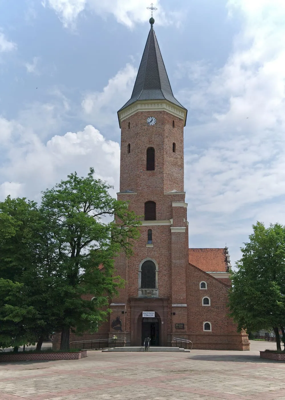 Ceglana wieża kościoła św. Mateusza w Pabianicach z ośmiobocznym zwieńczeniem i ciemnym, spiczastym hełmem, wśród zieleni
