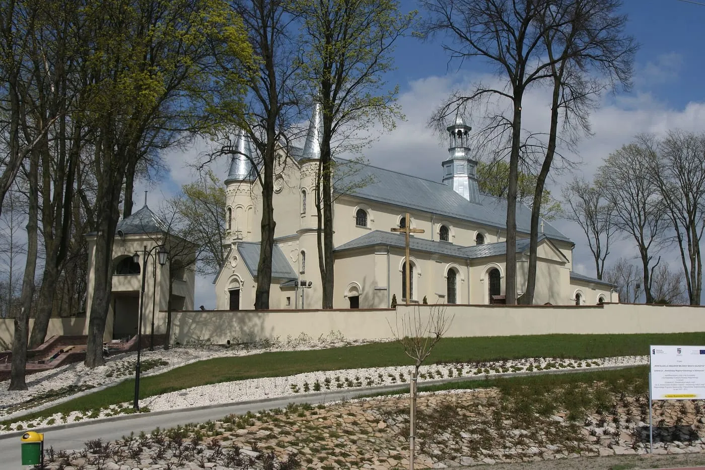 Kremowy kościół na wzgórzu z dwiema wieżyczkami o srebrnych hełmach i bramą-dzwonnicą