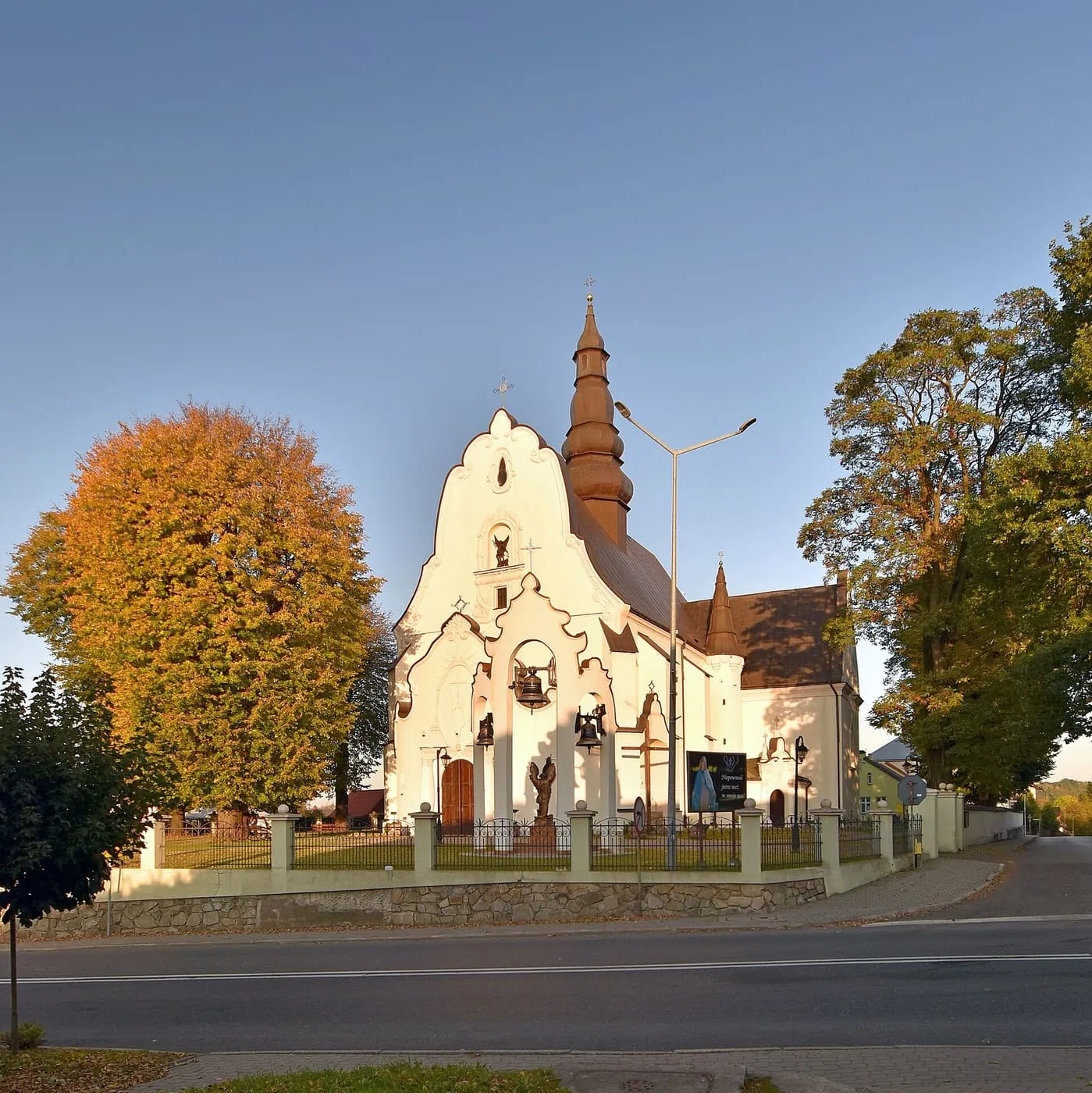 Biały kościół ze schodkowym szczytem i wieżyczką z cebulastym hełmem, otoczony drzewami o jesiennych barwach