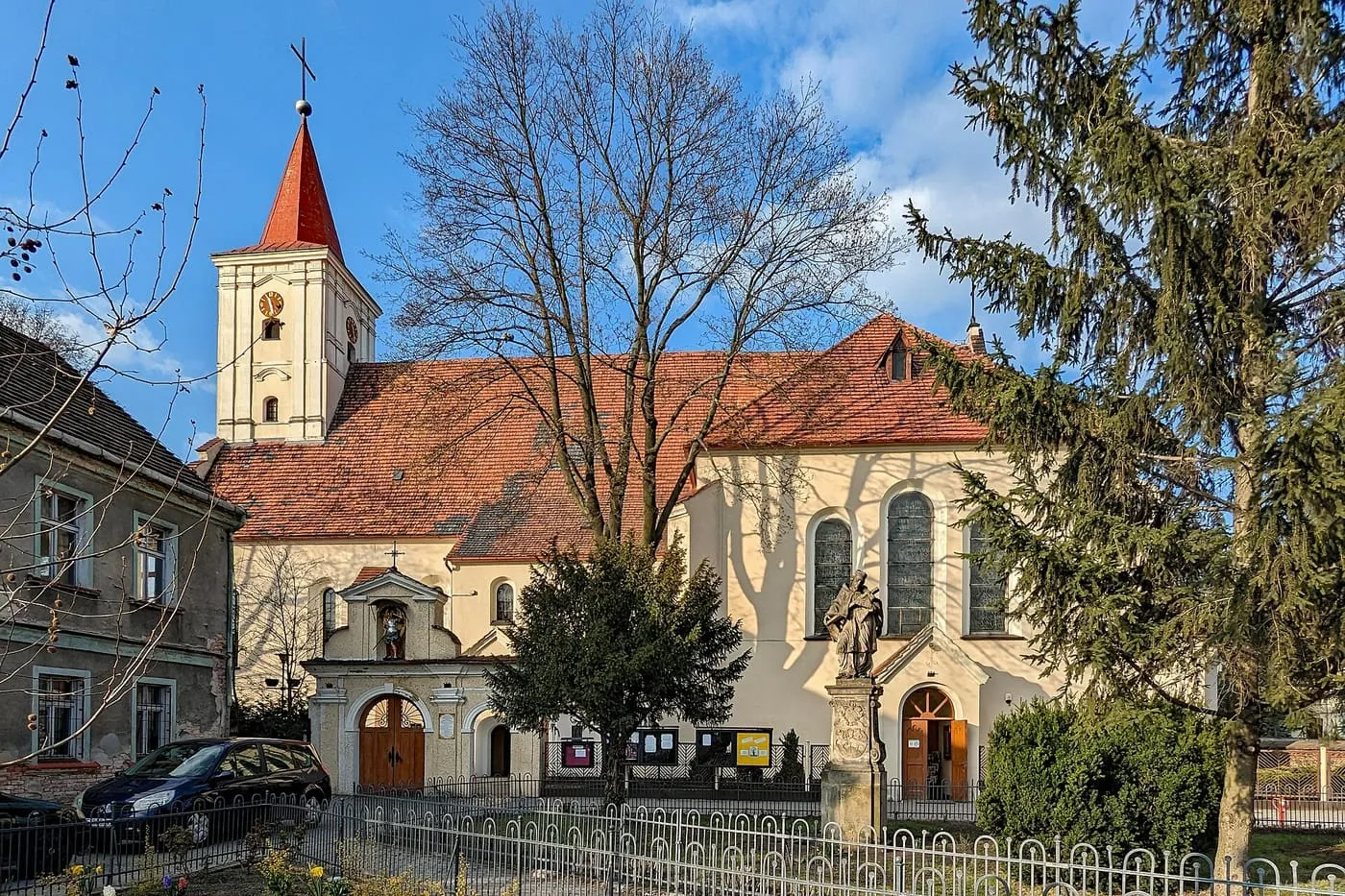 Kościół z jasną fasadą i czerwonym dachem, niską wieżą z zegarem i czerwonym hełmem, z barokowym portalem, ogrodzeniem i figurą przed wejściem, obok stoi świerk