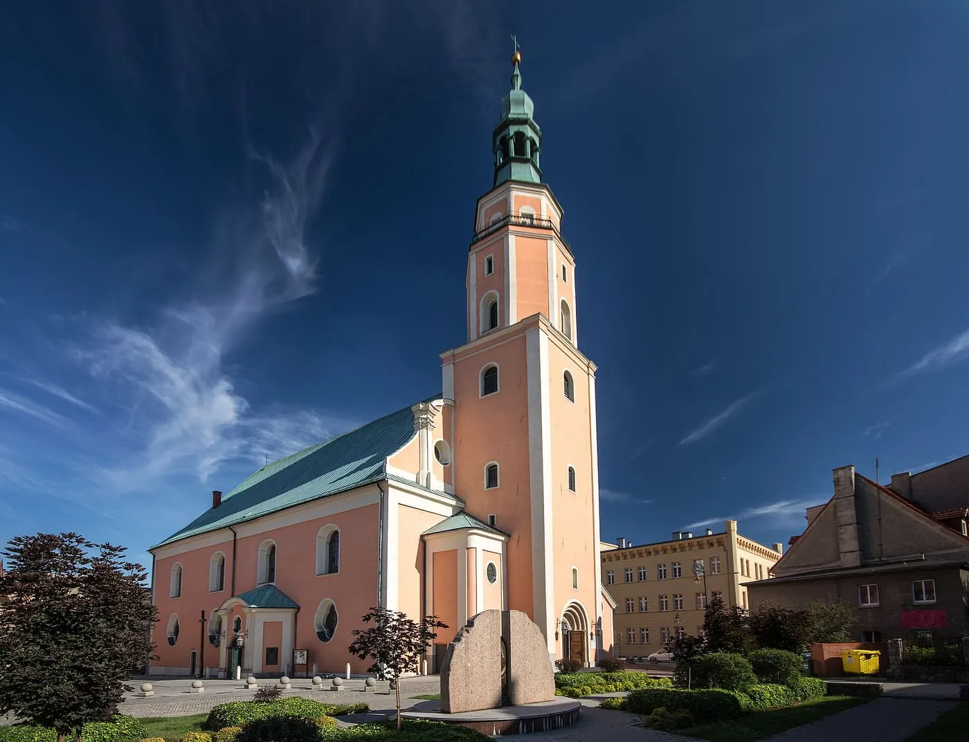 Łososiowa, barokowa fasada kościoła z wysoką wieżą z zielonym hełmem i zielonym dachem nawy, przy placu z głazem i klombami, pod ciemnoniebieskim niebem