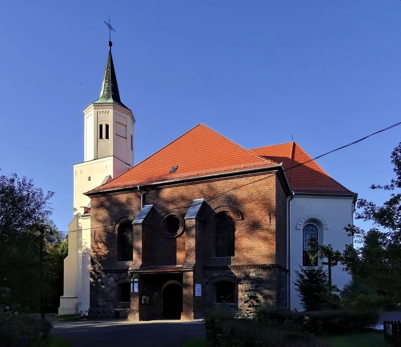 Kościół z ceglaną fasadą z okrągłym oknem i portalem, czerwonym dachem oraz jasną wieżą z ciemnym, ostrym hełmem na tle granatowego nieba