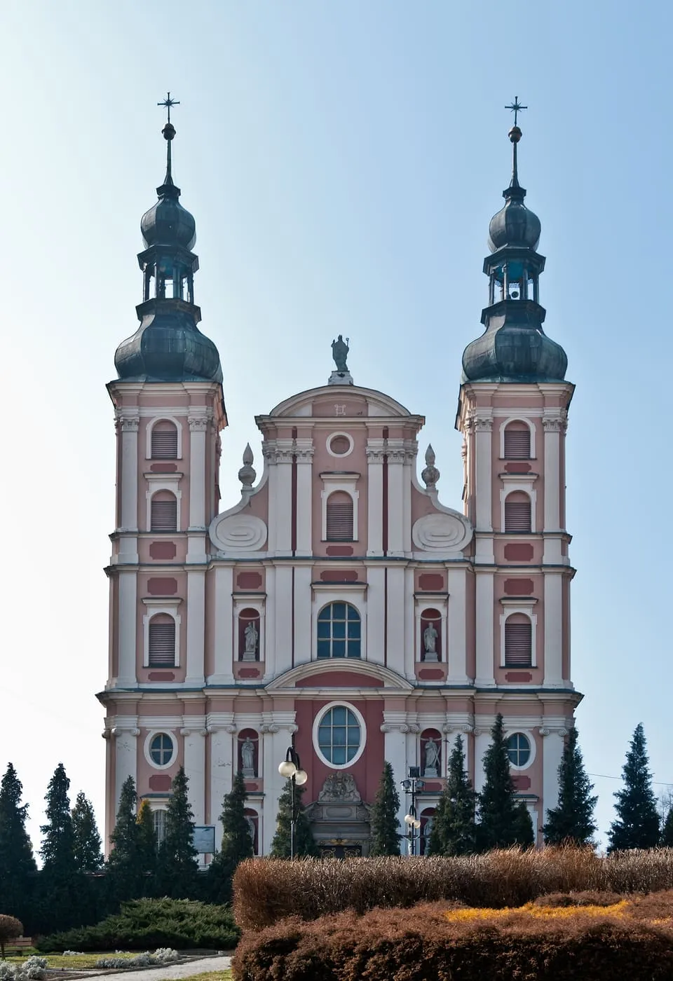 Barokowa fasada z dwiema smukłymi wieżami o zielonych hełmach, pomalowana na biało i różowo, za klombem i świerkami, na tle jasnego nieba