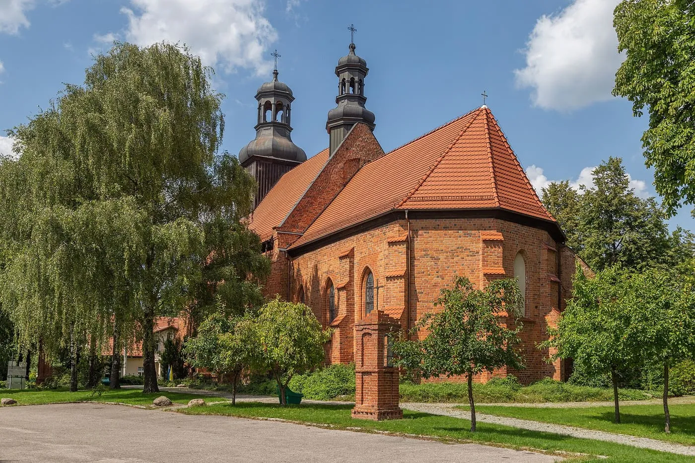 Ceglany, gotycki kościół ze stromym, czerwonym dachem i drewnianą wieżą zwieńczoną ciemnymi, barokowymi hełmami, z płaczącymi wierzbami na pierwszym planie