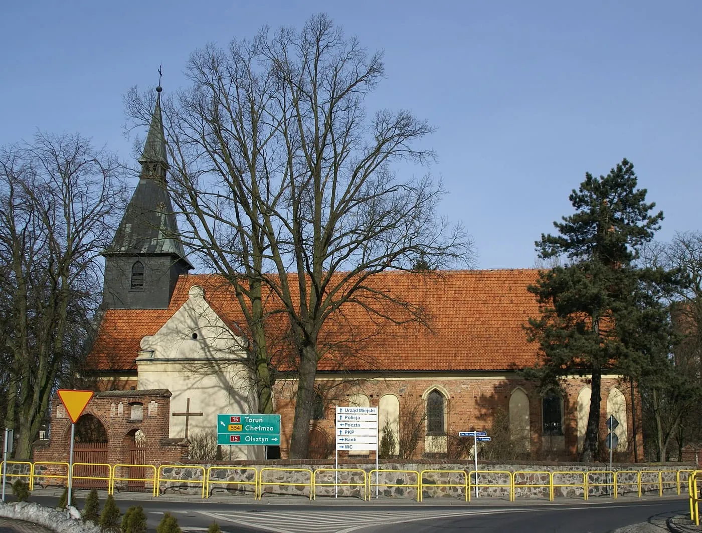 Gotycki kościół z cegły i kamienia z czerwonym dachem, niską wieżą z ciemnym, spiczastym hełmem i jasnym szczytem, widziany zza bezlistnych drzew i żółtej barierki przy skrzyżowaniu