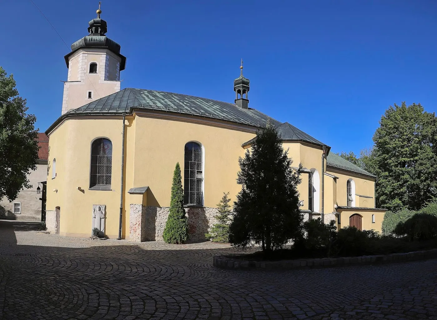 Żółty kościół z zielonym dachem, niską wieżą z ciemnym hełmem i wieżyczką na sygnaturkę, przy brukowanym placu, na tle intensywnie błękitnego nieba