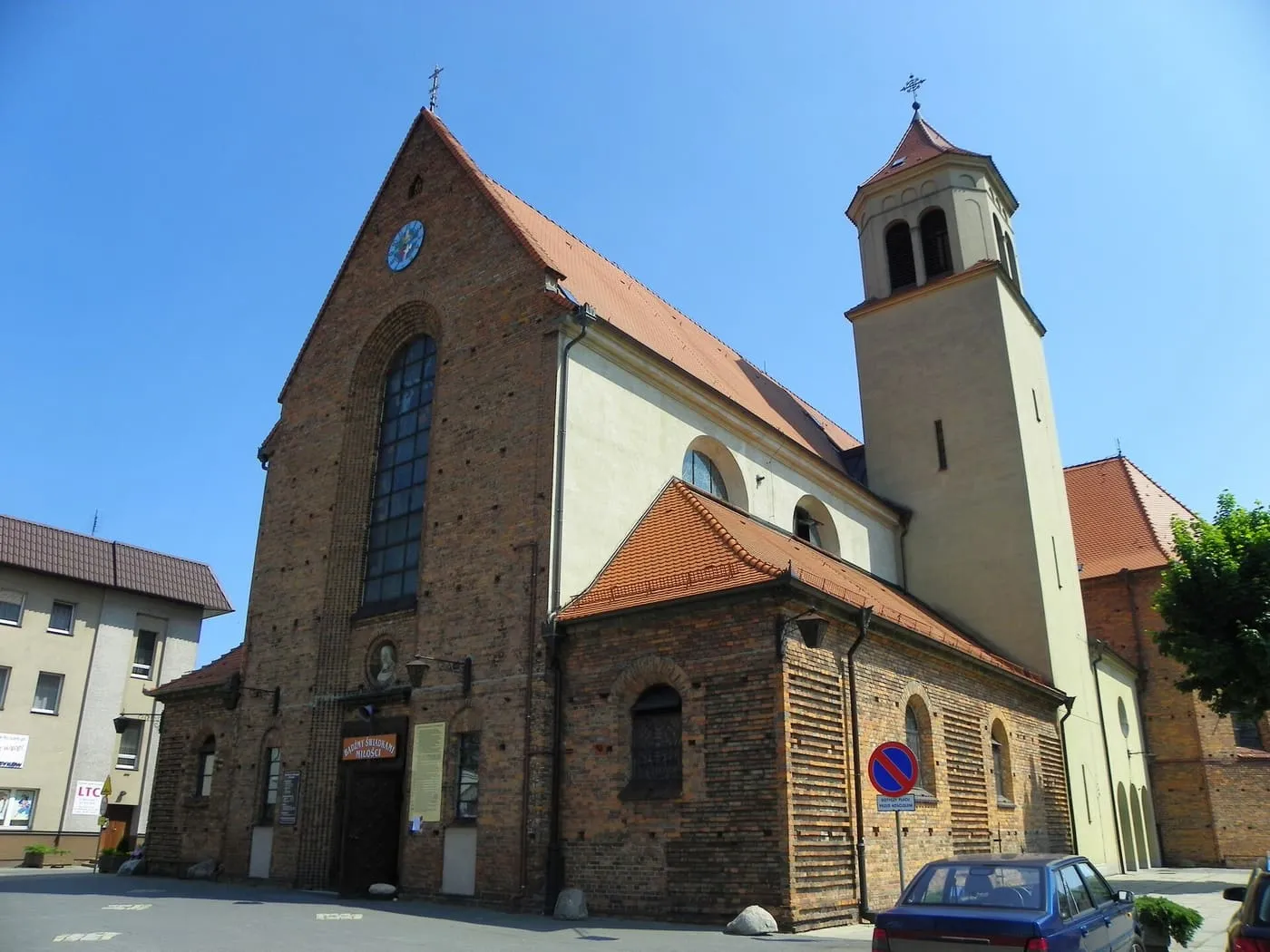 Ceglany kościół z wysoką, jasną wieżą z zegarem, dużym oknem w ceglanej fasadzie i czerwonymi dachami, przy ulicy z zaparkowanym samochodem
