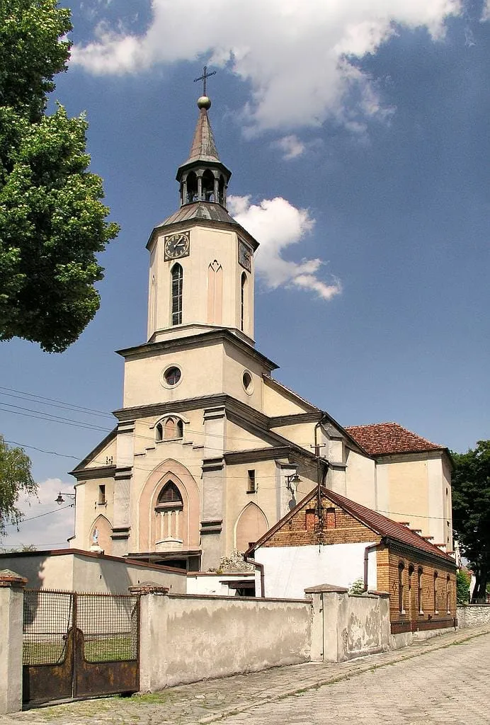 Kremowy kościół z wieżą zegarową i latarnią, przy nim mur z bramą i brukowana ulica