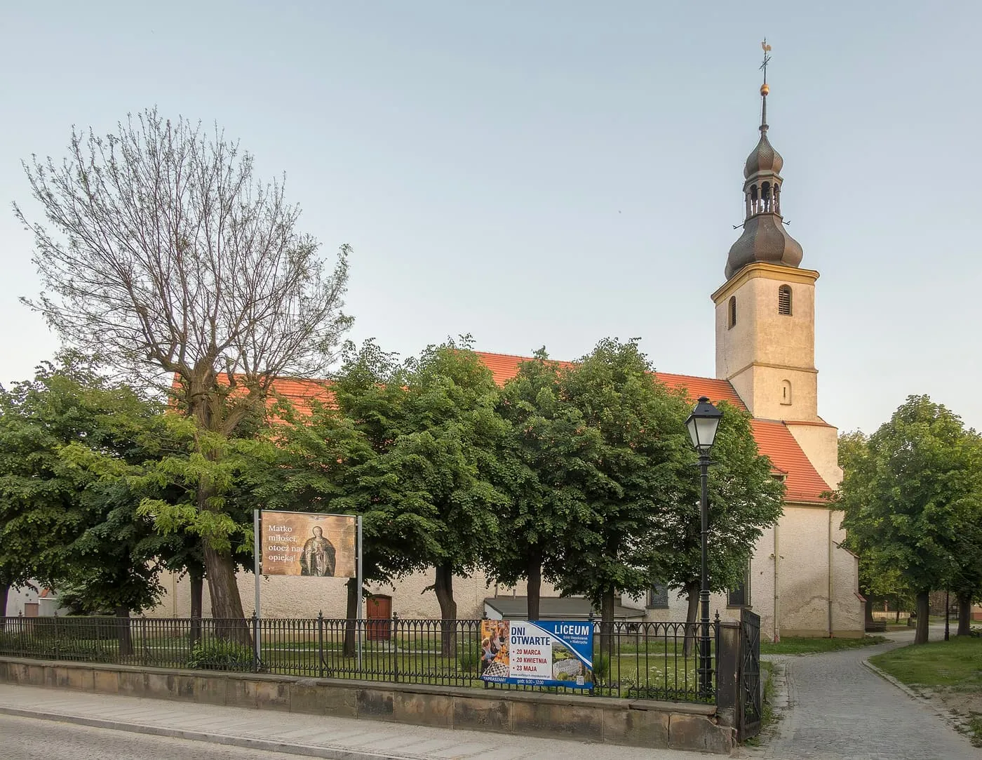Kamienny kościół z czerwonym dachem i żółtą wieżą z ciemnym, barokowym hełmem, widziany zza drzew i ulicznego ogrodzenia, na tle bladego nieba