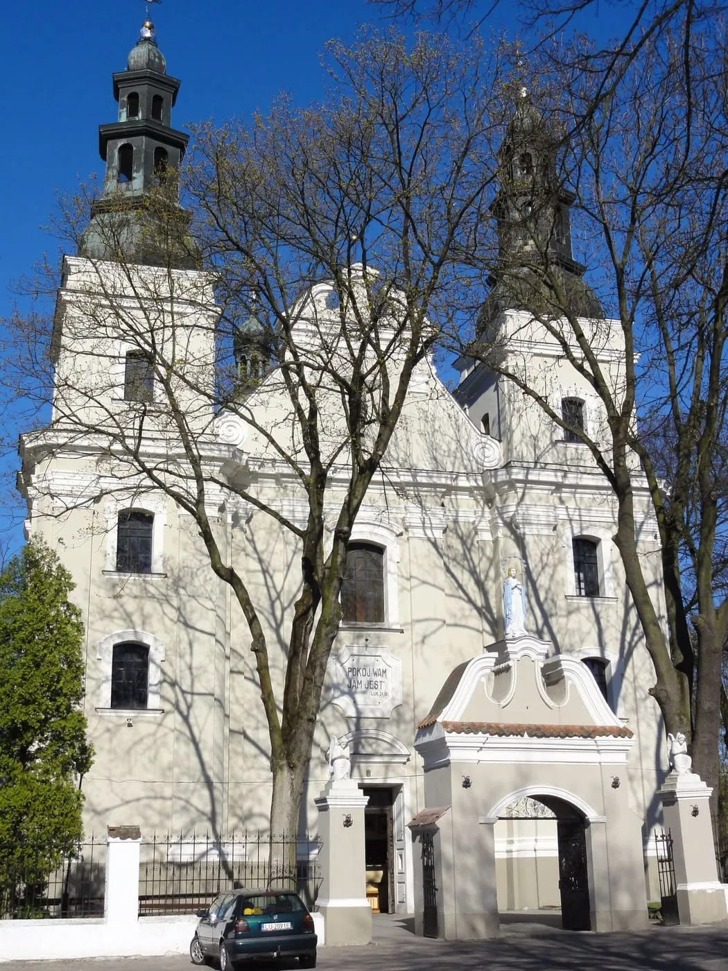 Biała, dwuwieżowa fasada barokowego kościoła z ciemnymi hełmami wież, widziana zza bezlistnych drzew; przed wejściem jasna brama, na pierwszym planie samochód