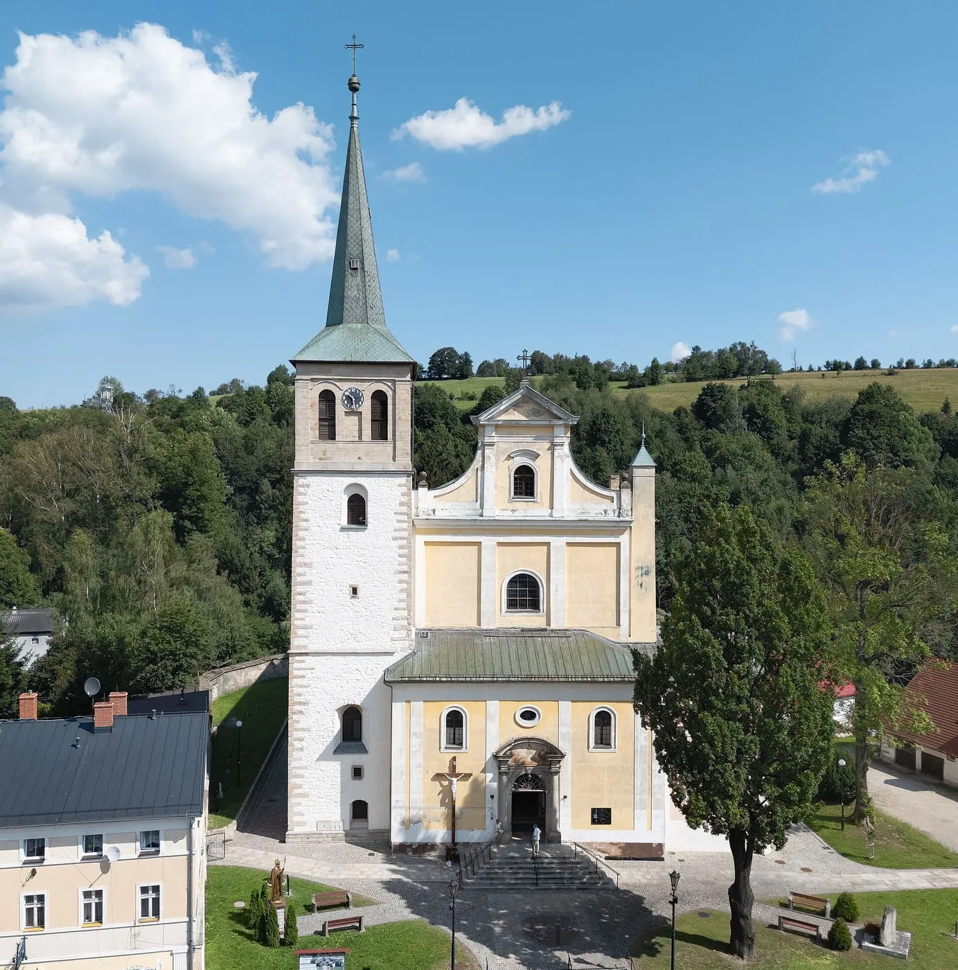 Żółty, barokowy kościół z białą wieżą zwieńczoną wysoką, zieloną iglicą, ze schodami do wejścia, na tle zalesionego wzgórza
