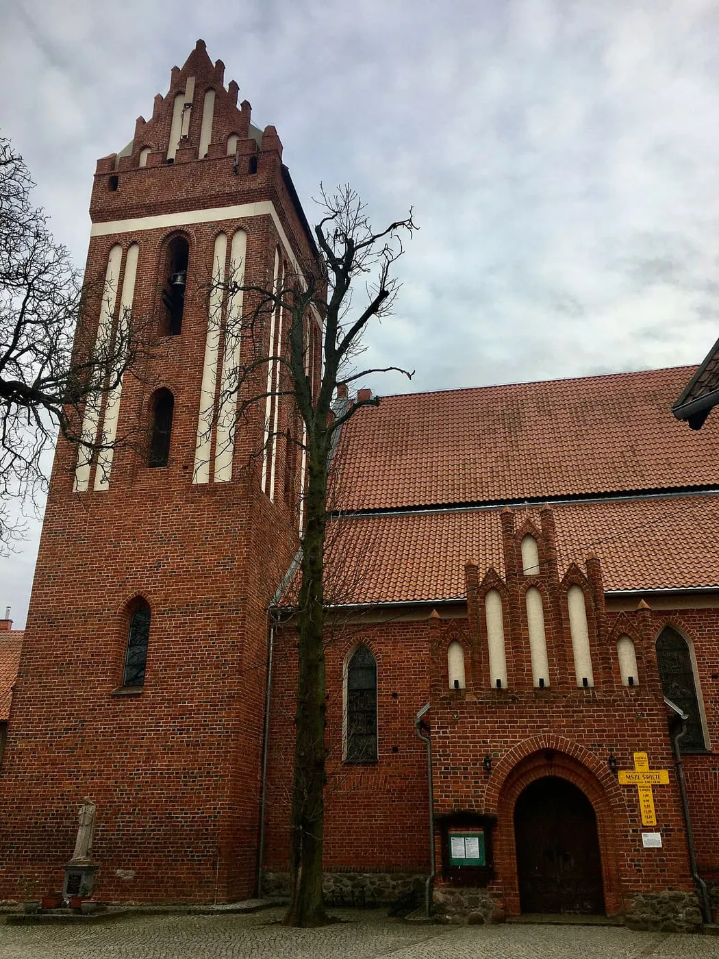 Gotycki kościół z czerwonej cegły z wysoką wieżą z jasnymi wnękami i schodkowym szczytem, przy niej portal z kruchtą; na pierwszym planie bezlistne drzewo