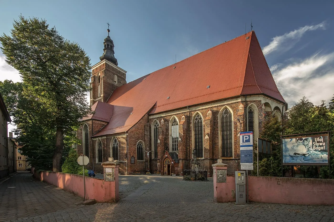 Gotycki, ceglany kościół z wysokim czerwonym dachem i kwadratową wieżą, widziany z brukowanego placu i różowego muru, w wieczornym świetle