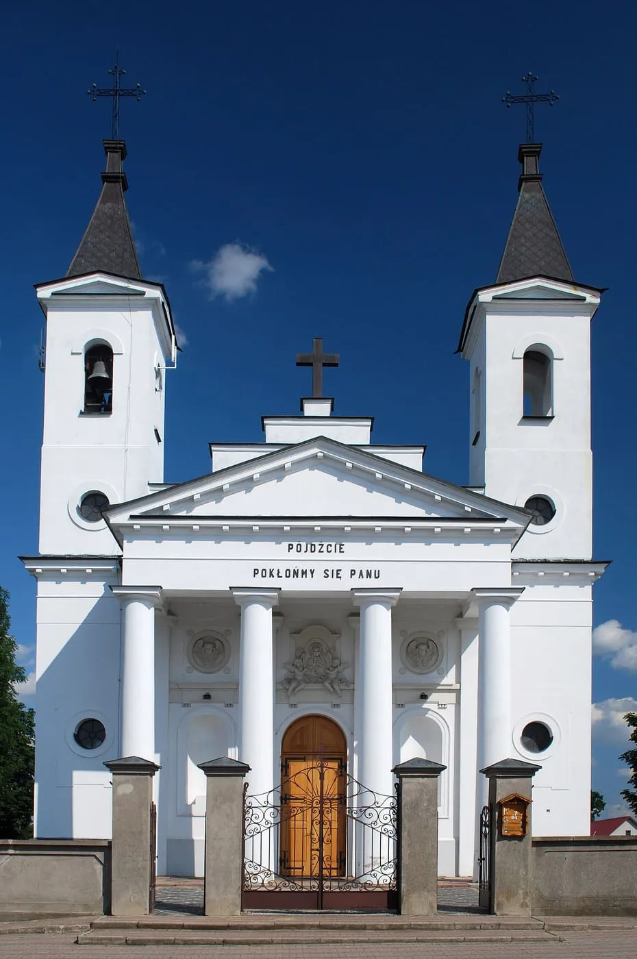 Klasycystyczna, biała fasada kościoła w Zabłudowie z dwiema symetrycznymi wieżami zwieńczonymi krzyżami, kolumnowym portykiem i drewnianymi drzwiami za kutą bramą, na tle granatowego nieba