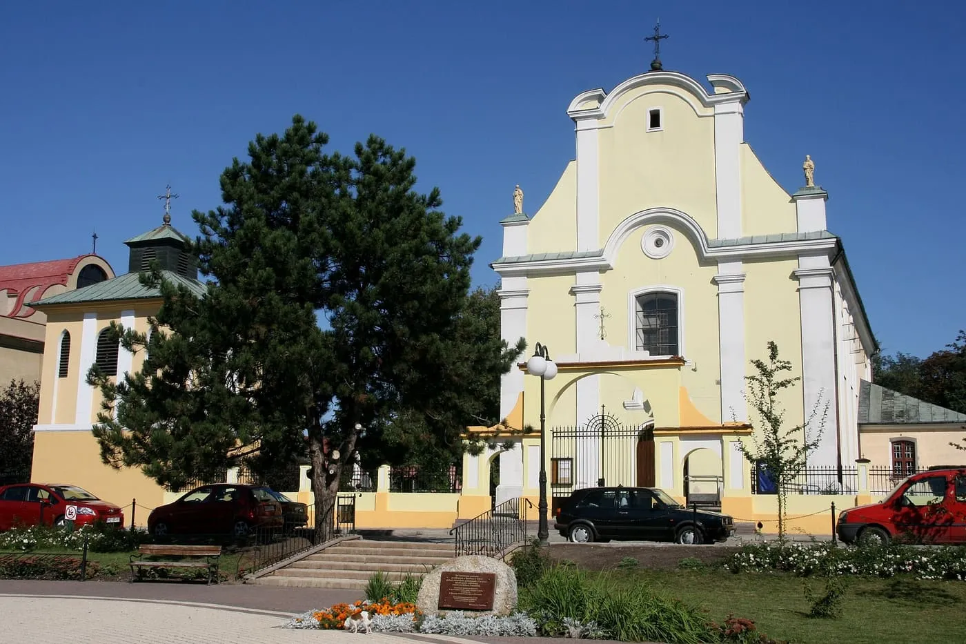 Kremowo-żółty, barokowy kościół z falistym szczytem fasady i żelazną bramą przed wejściem; z lewej drzewo i kaplica z zielonym dachem, na pierwszym planie skwer i samochody