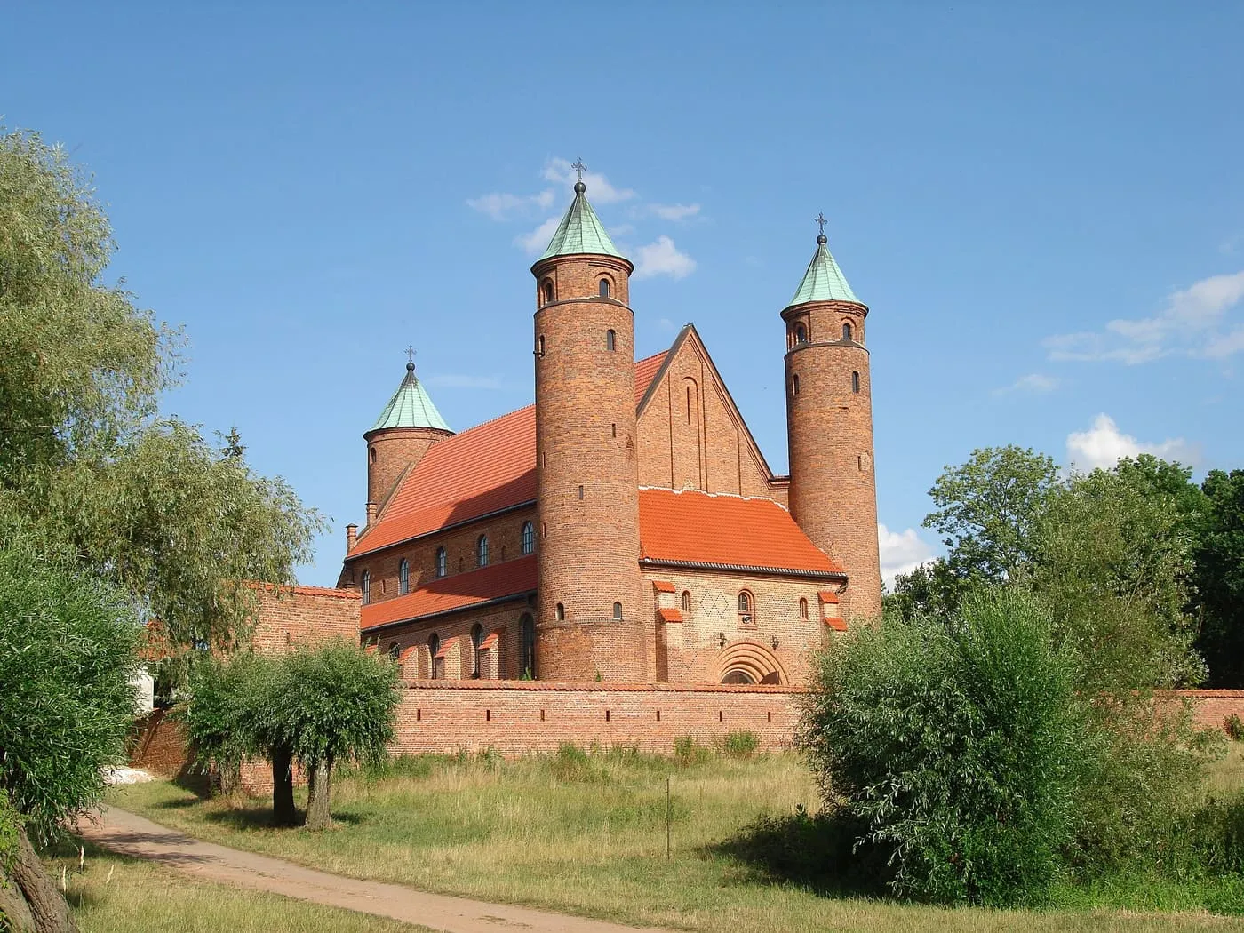 Ceglany kościół w Brochowie z trzema cylindrycznymi wieżami nakrytymi zielonymi, stożkowymi hełmami, otoczony ceglanym murem, widziany od strony łąki