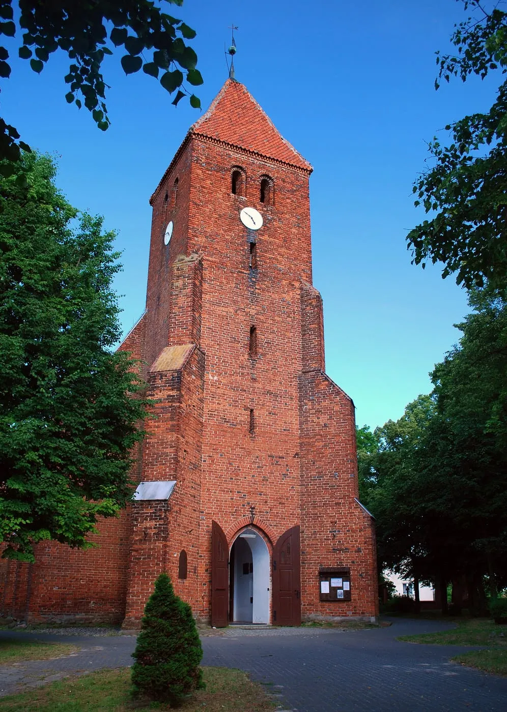 Ceglana, gotycka wieża kościoła z niewielką iglicą, otoczona zielenią, z widocznym wejściem u dołu