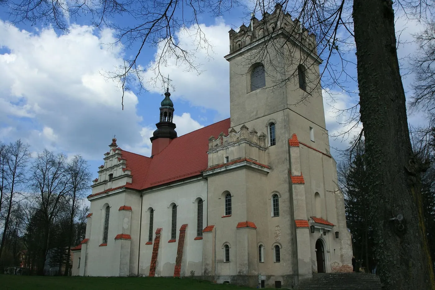 Bielony kościół z wieżą zwieńczoną attyką z krenelażem i czerwonym dachem z dachówki, wśród bezlistnych drzew