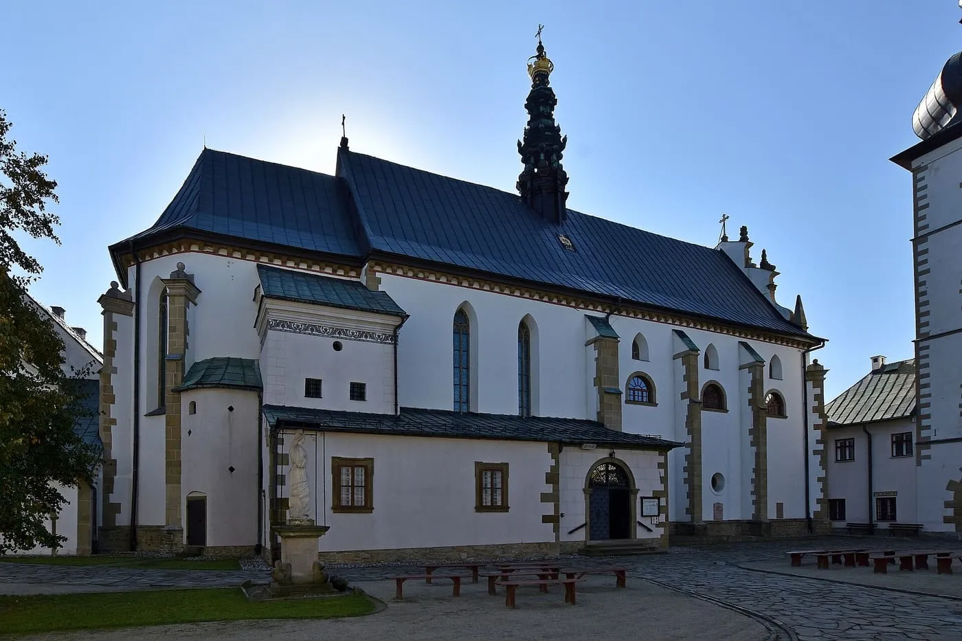 Bielony kościół klarysek w Starym Sączu z ciemnym dachem i ażurową, czarno-złotą wieżyczką na sygnaturkę zwieńczoną koroną, widziany z dziedzińca klasztornego