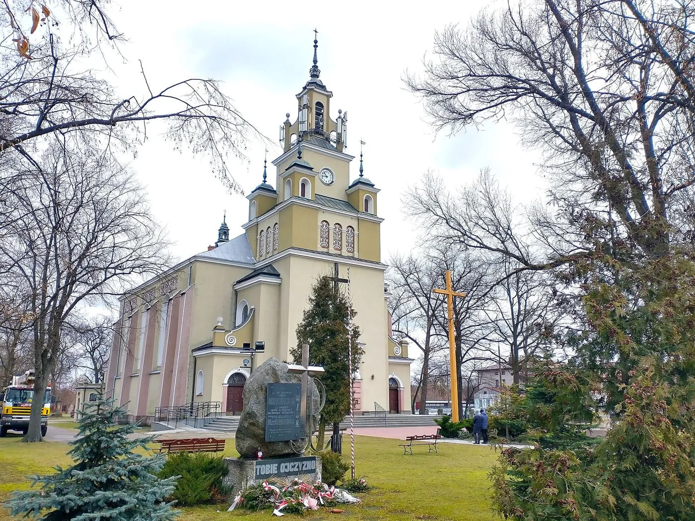 Żółty kościół z wieżą z zegarem i sygnaturkami, na pierwszym planie kamień pamiątkowy i krzyż, wśród gołych drzew na trawniku