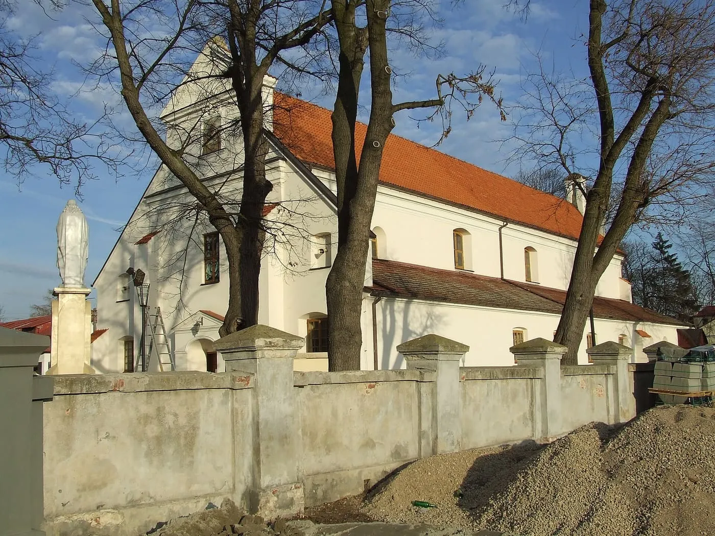 Bielony kościół Świętej Trójcy w Błoniu z czerwonym dachem i schodkowym szczytem, widziany zza muru cmentarza przykościelnego z białą figurą Matki Bożej na cokole