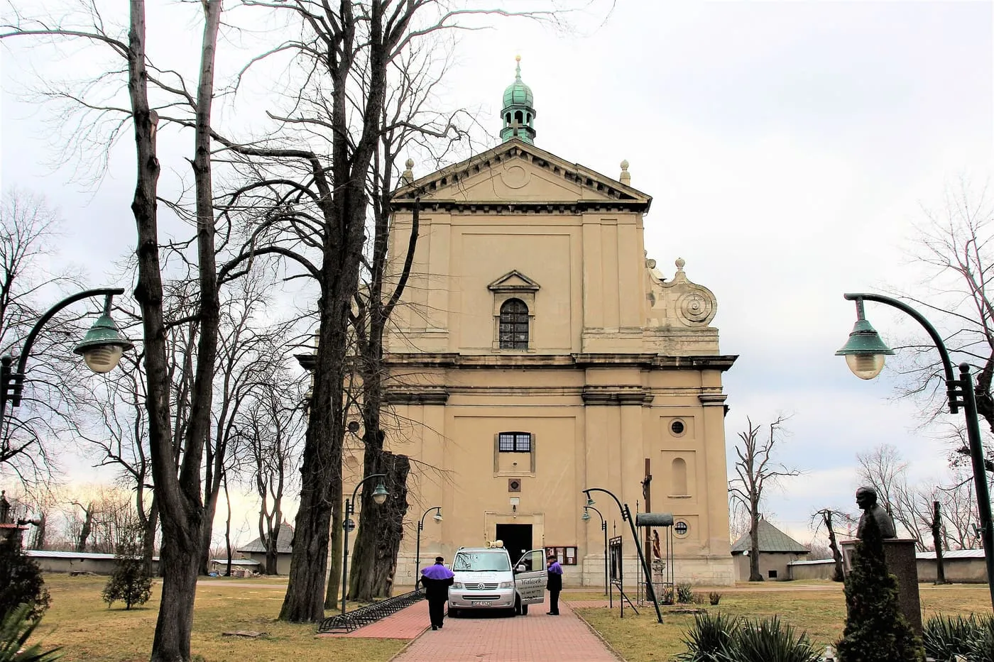 Barokowa, beżowa fasada kościoła z trójkątnym frontonem i zielonym hełmem wieżyczki, do której prowadzi ceglana alejka wśród gołych drzew, z latarniami po bokach
