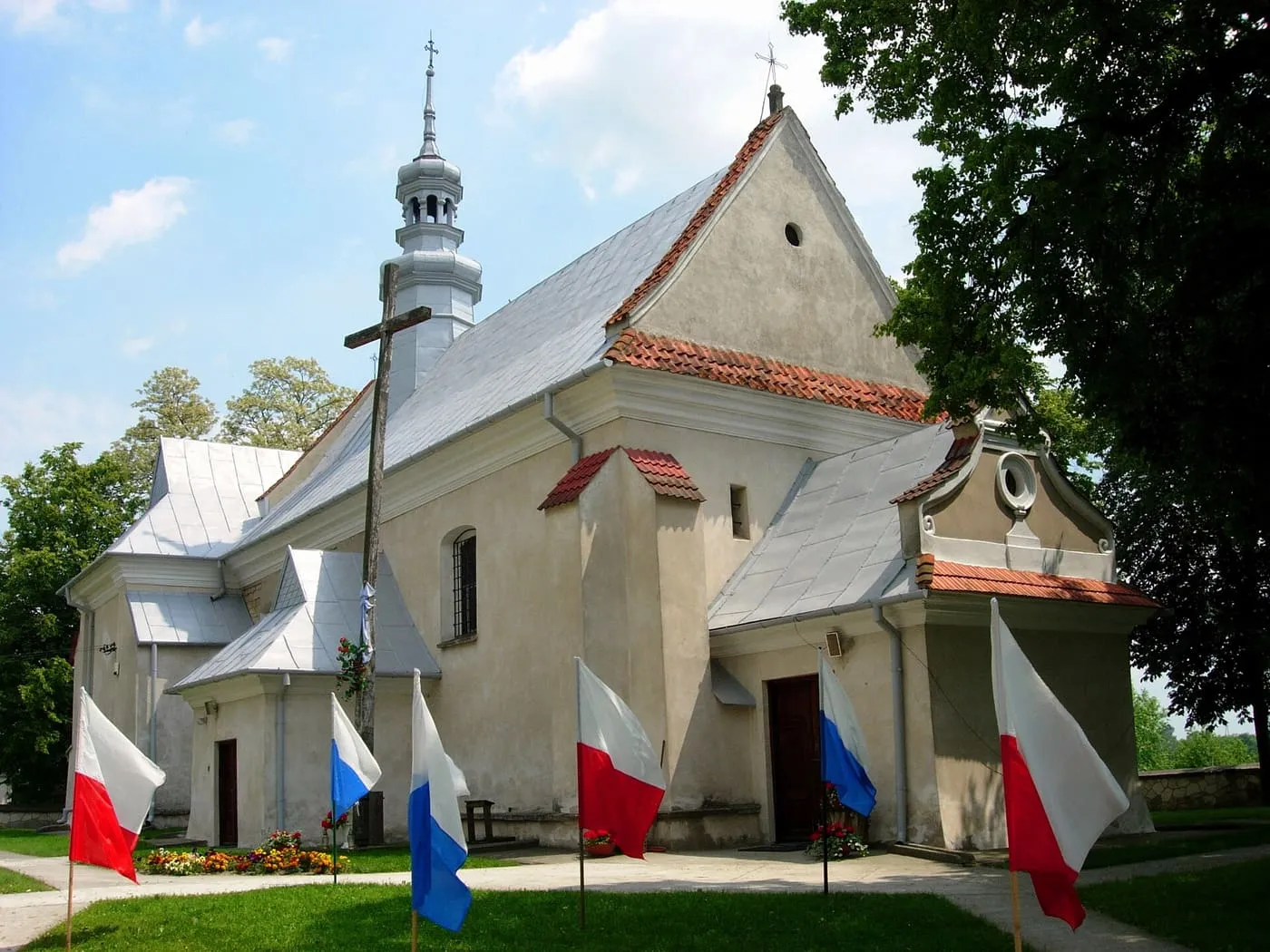 Biały kościół z ceglastą dachówką na pochyłościach dachu i wieżyczką na sygnaturkę, przed nim na trawniku biało-czerwone i biało-niebieskie flagi, w tle wysokie drzewa