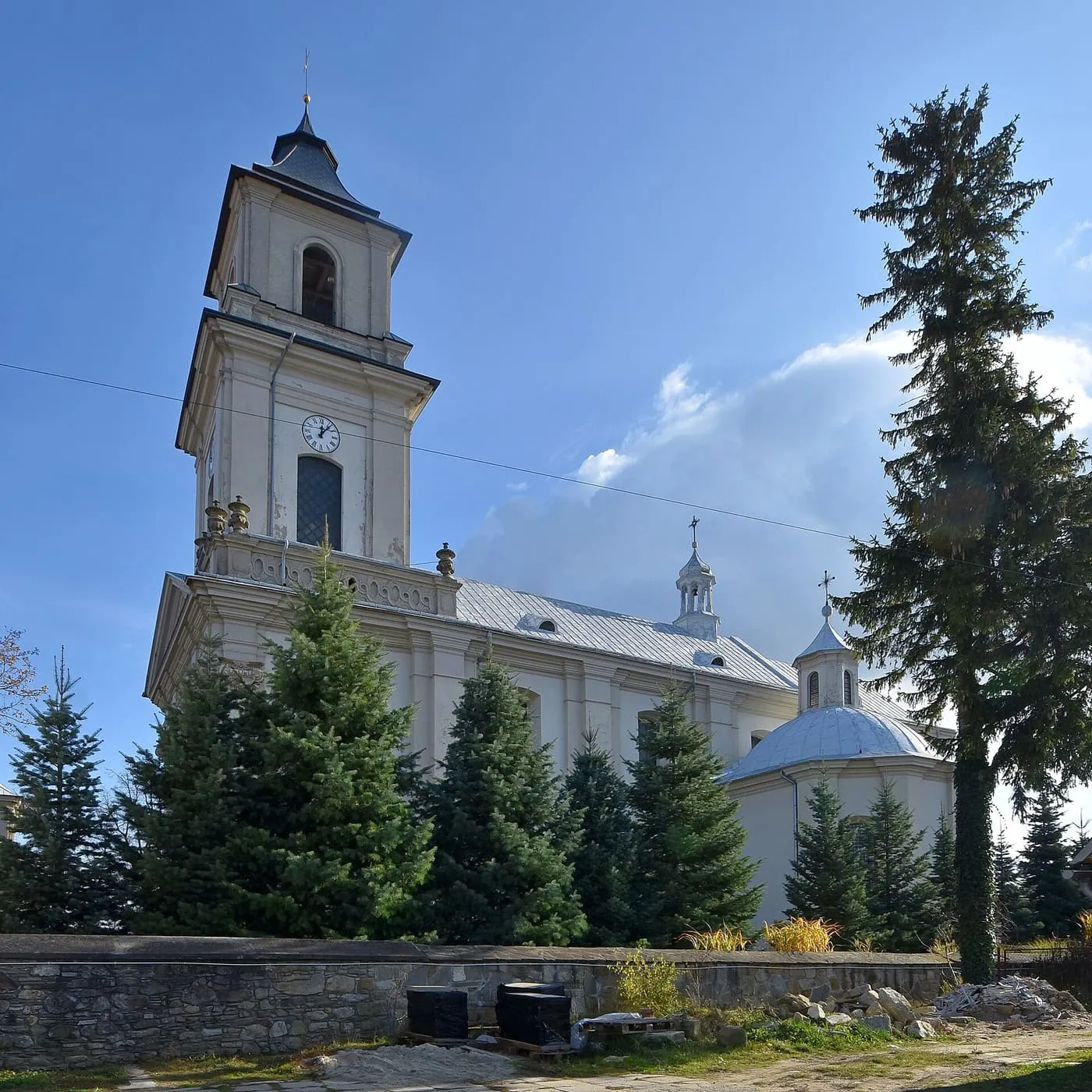 Biały, barokowy kościół z wysoką wieżą z zegarem i kopułą kaplicy z boku; przed nim rosną świerki, a niżej ciągnie się kamienny mur