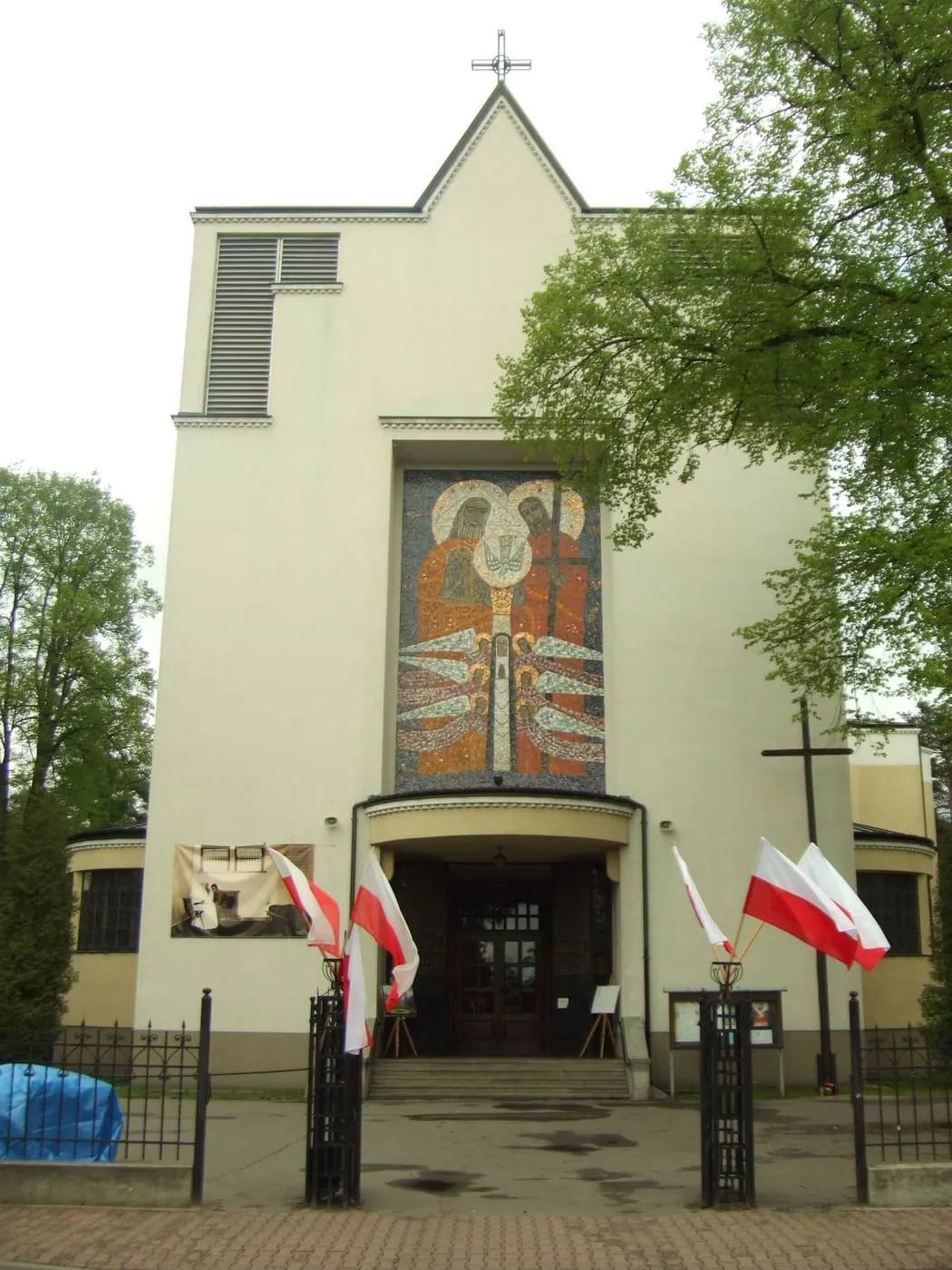 Modernistyczna fasada kościoła z dużym mozaikowym wizerunkiem Matki Bożej nad wejściem i polskimi flagami