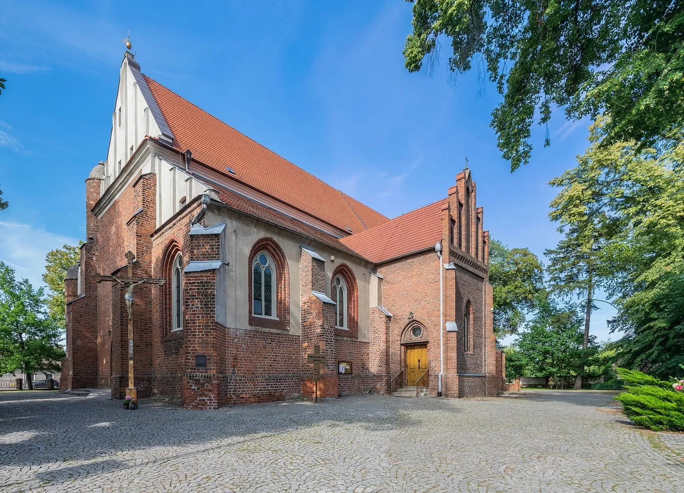 Ceglany, gotycki kościół z czerwonym dachem i schodkowym szczytem kruchty, z drewnianym krzyżem przy ścianie i brukowanym placem przed wejściem, w słońcu, pod błękitnym niebem