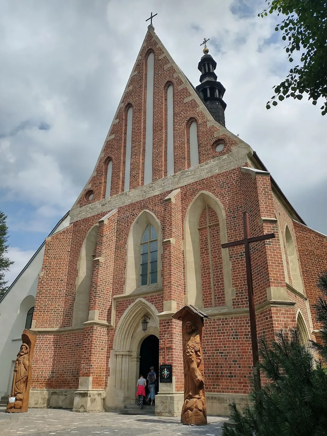Ceglany, gotycki szczyt kościoła św. Władysława w Szydłowie z wąskimi, ostrołukowymi oknami, drewnianą dzwonnicą w tle i rzeźbionymi figurami świętych przy wejściu