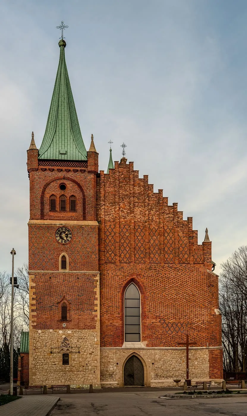 Gotycki kościół z czerwonej cegły z wysoką wieżą z zegarem i zielonym, ostrym hełmem oraz schodkowym szczytem nawy; u dołu kamienna podmurówka i ostrołukowy portal