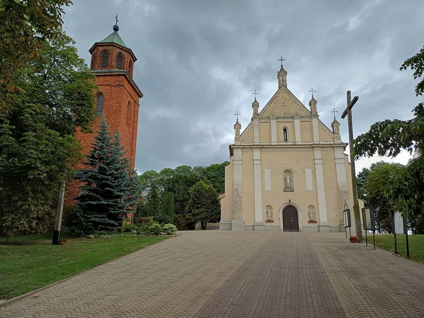 Jasna, barokowa fasada kościoła z trójkątnym szczytem i wieżyczkami, obok ceglana dzwonnica, do których prowadzi szeroki brukowany plac, pod pochmurnym niebem