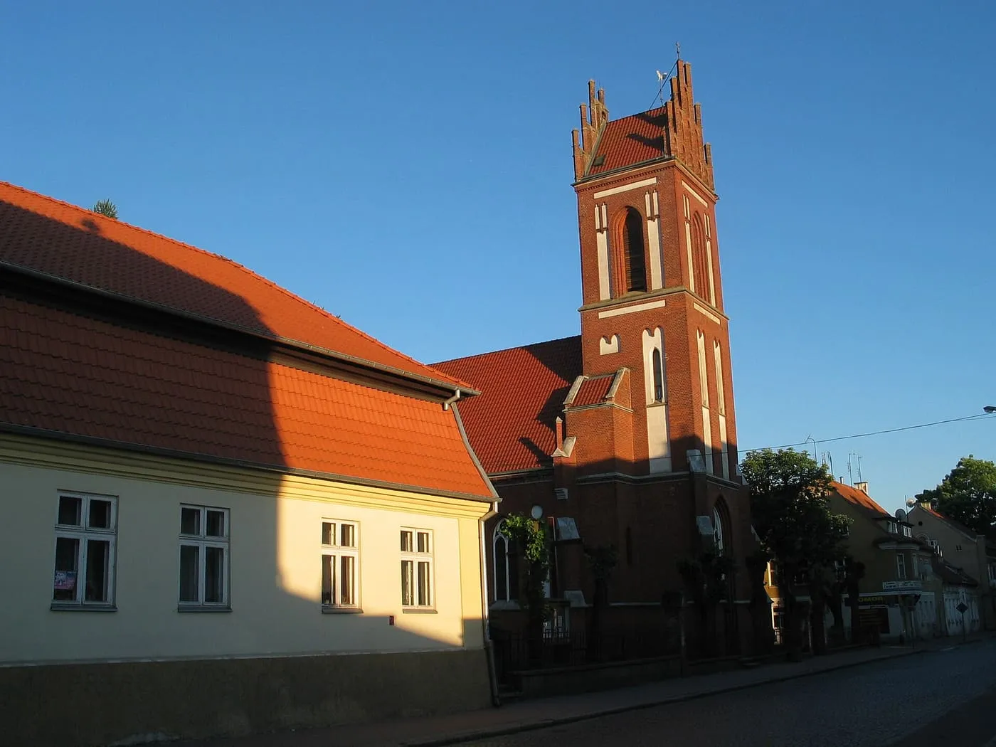 Ceglana wieża kościoła ze schodkowym szczytem i czerwonym dachem wznosi się za żółtym budynkiem przy brukowanej ulicy, w ciepłym świetle zachodu słońca