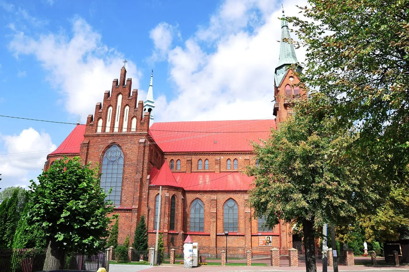 Neogotycki kościół z czerwonej cegły, ze schodkowym szczytem z wysokim, ostrołukowym oknem, czerwonym dachem i smukłą wieżą z zielonym hełmem, wśród drzew
