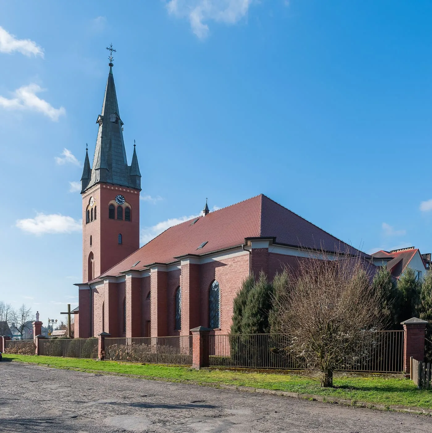 Ceglany, neogotycki kościół z wysoką wieżą z zegarem i ciemnym hełmem, widziany z boku zza ogrodzenia, na tle błękitnego nieba