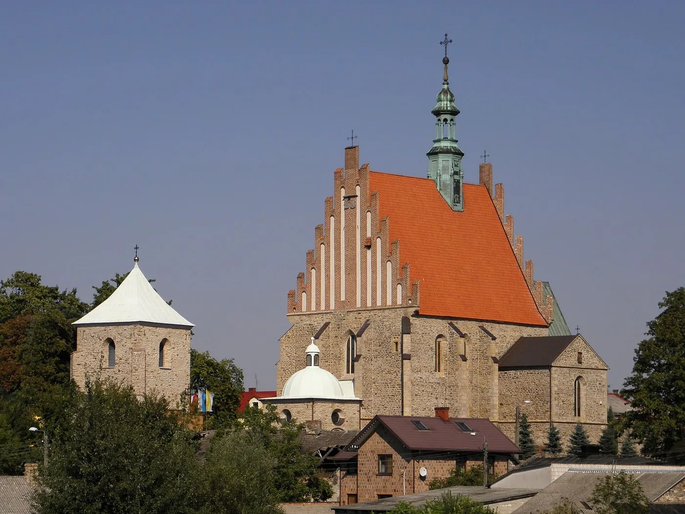 Kamienna fara z ceglanym, schodkowym szczytem i pomarańczowym dachem, ze zieloną sygnaturką; po lewej murowana dzwonnica z białym dachem, u dołu kopulasta kaplica
