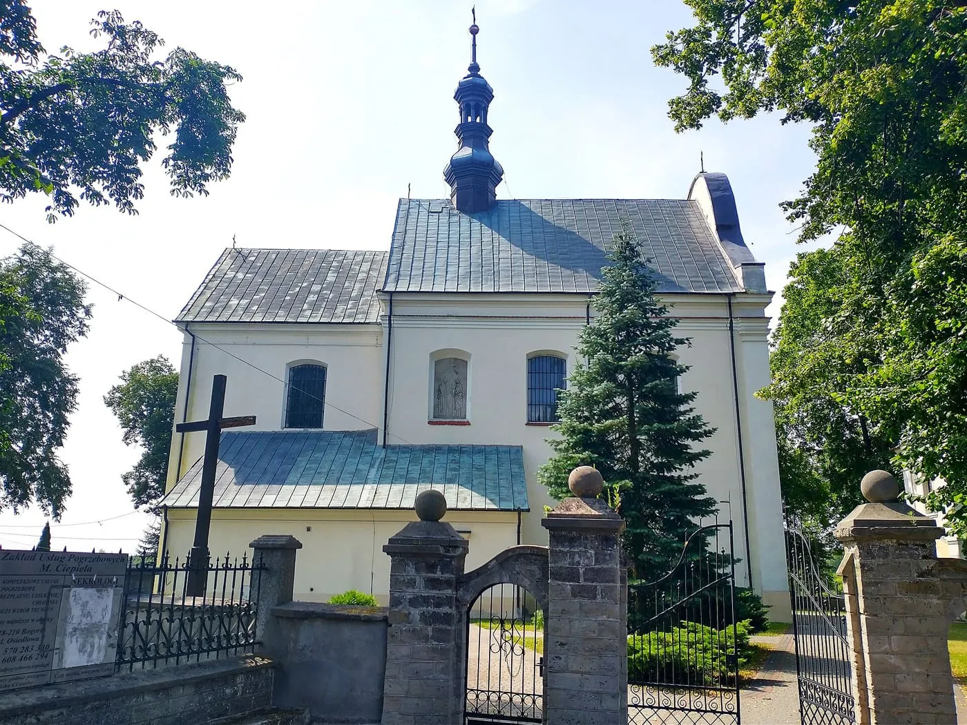 Biały kościół z blaszanym dachem i wieżyczką z ciemnym, cebulastym hełmem, widziany zza kamiennej bramy z kutymi skrzydłami i drewnianego krzyża, wśród drzew