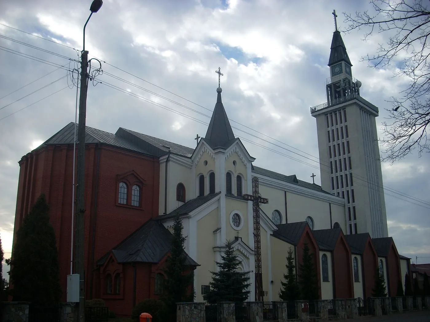 Kościół z jasną fasadą o spiczastym szczycie, czerwoną ceglaną bryłą prezbiterium i wysoką, smukłą wieżą dzwonną, widziany z ukosa spod pochmurnego nieba