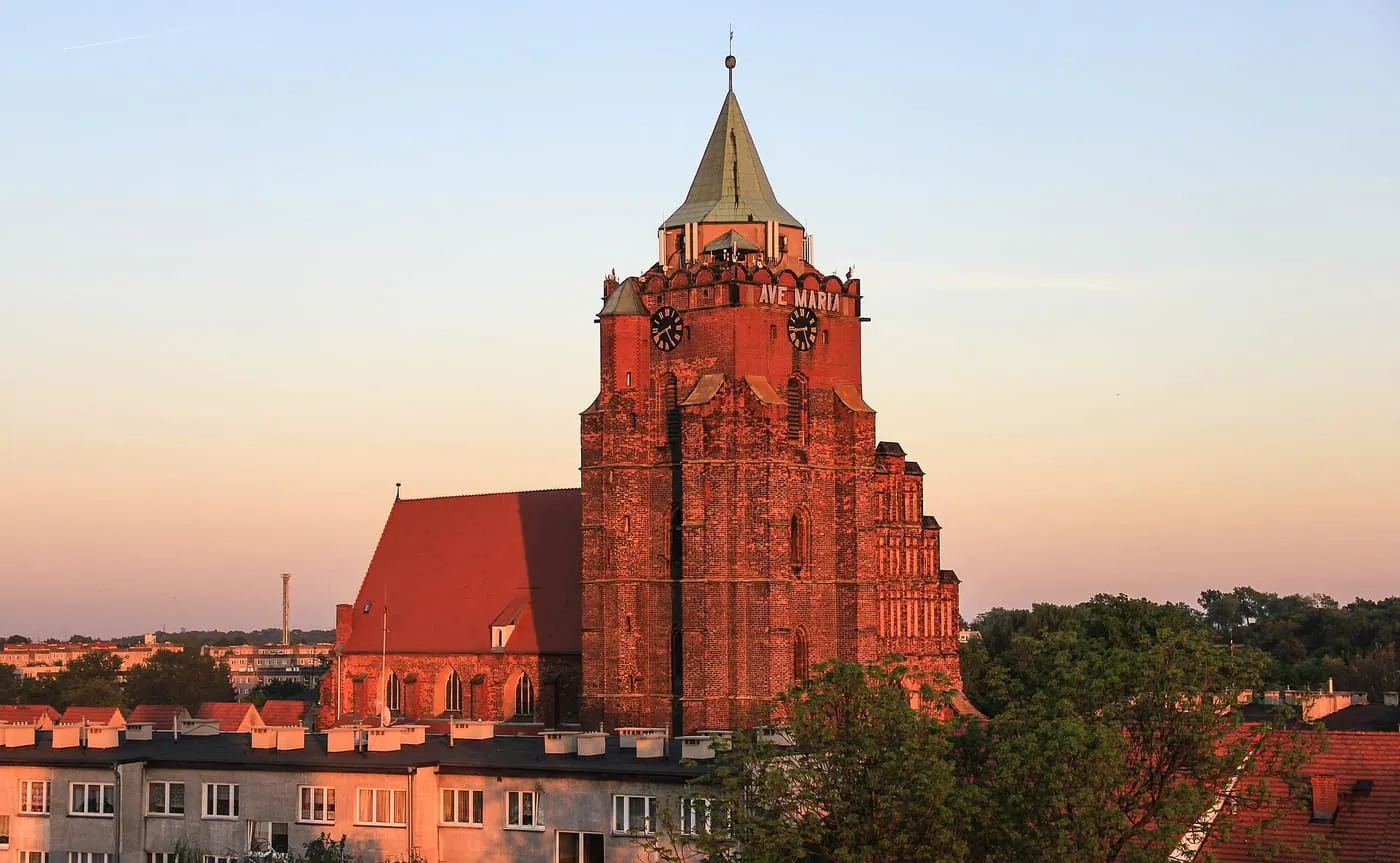 Masywna ceglana wieża z krenelażem i stożkowym hełmem oraz stromy dach nawy, w zachodzącym słońcu nad dachami miasta