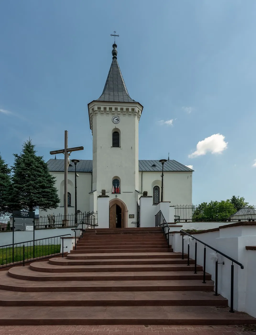 Biały kościół z wieżą z zegarem i cebulastym hełmem, u podnóża szerokie schody i krzyż, na tle błękitnego nieba
