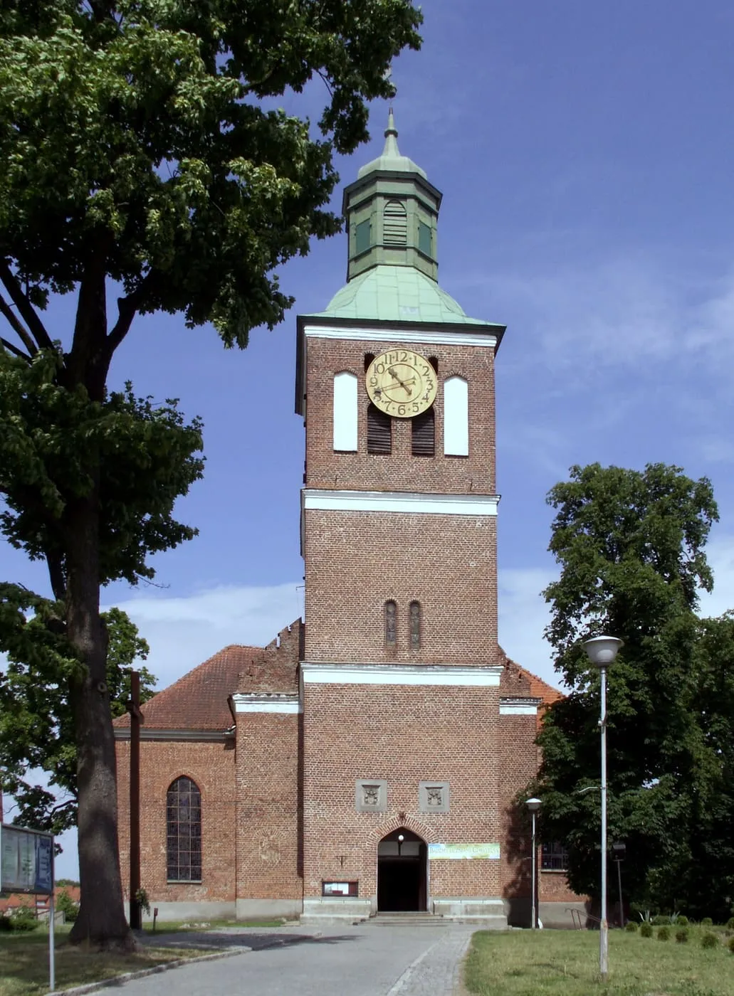 Ceglana wieża z zegarem, zwieńczona zielonym hełmem z latarnią, wznosi się nad frontem kościoła, obok rośnie duże drzewo, w tle niebo z chmurami