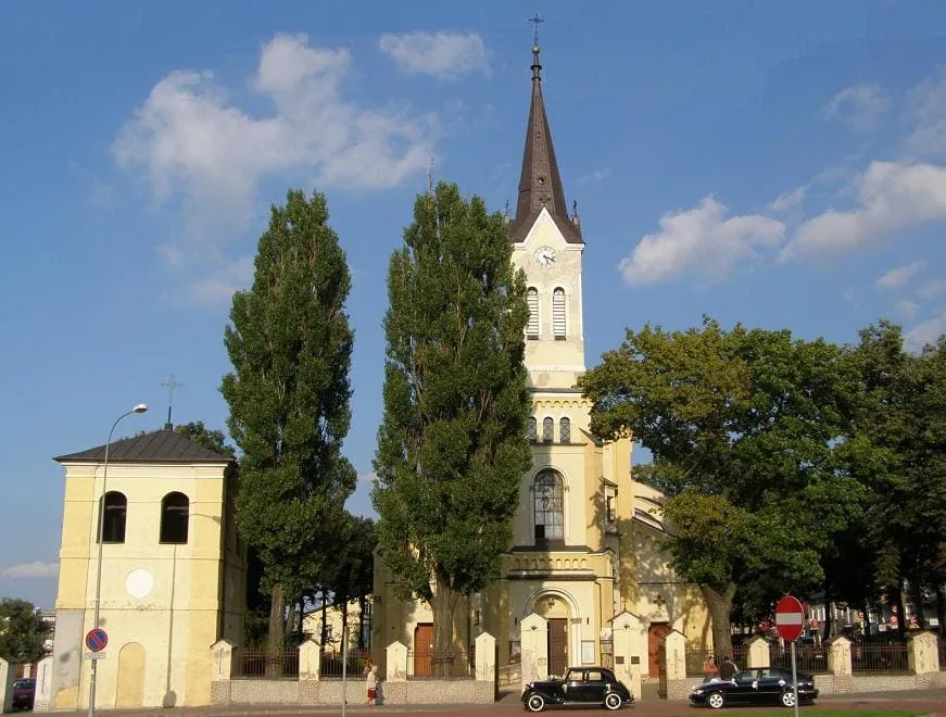 Kremowy kościół w Grajewie z pojedynczą wieżą z zegarem i strzelistym hełmem, otoczony wysokimi topolami, z osobno stojącą dzwonnicą i zabytkowym samochodem zaparkowanym przed wejściem