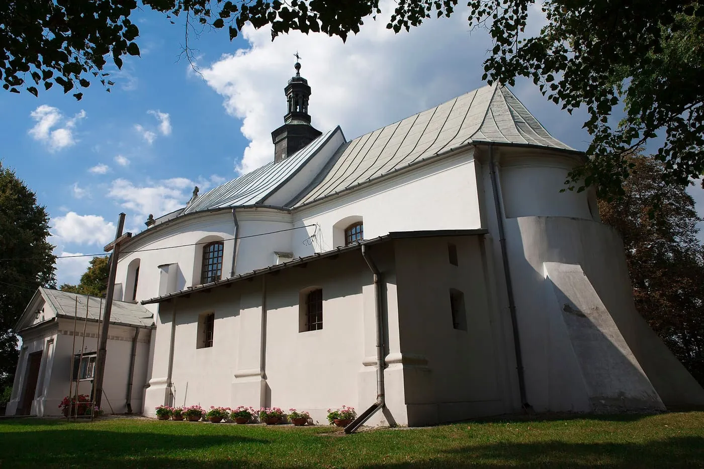 Biały, romańsko-barokowy kościół z szarym blaszanym dachem i małą wieżyczką, widziany z ukosa spod gałęzi drzew, na tle błękitnego nieba