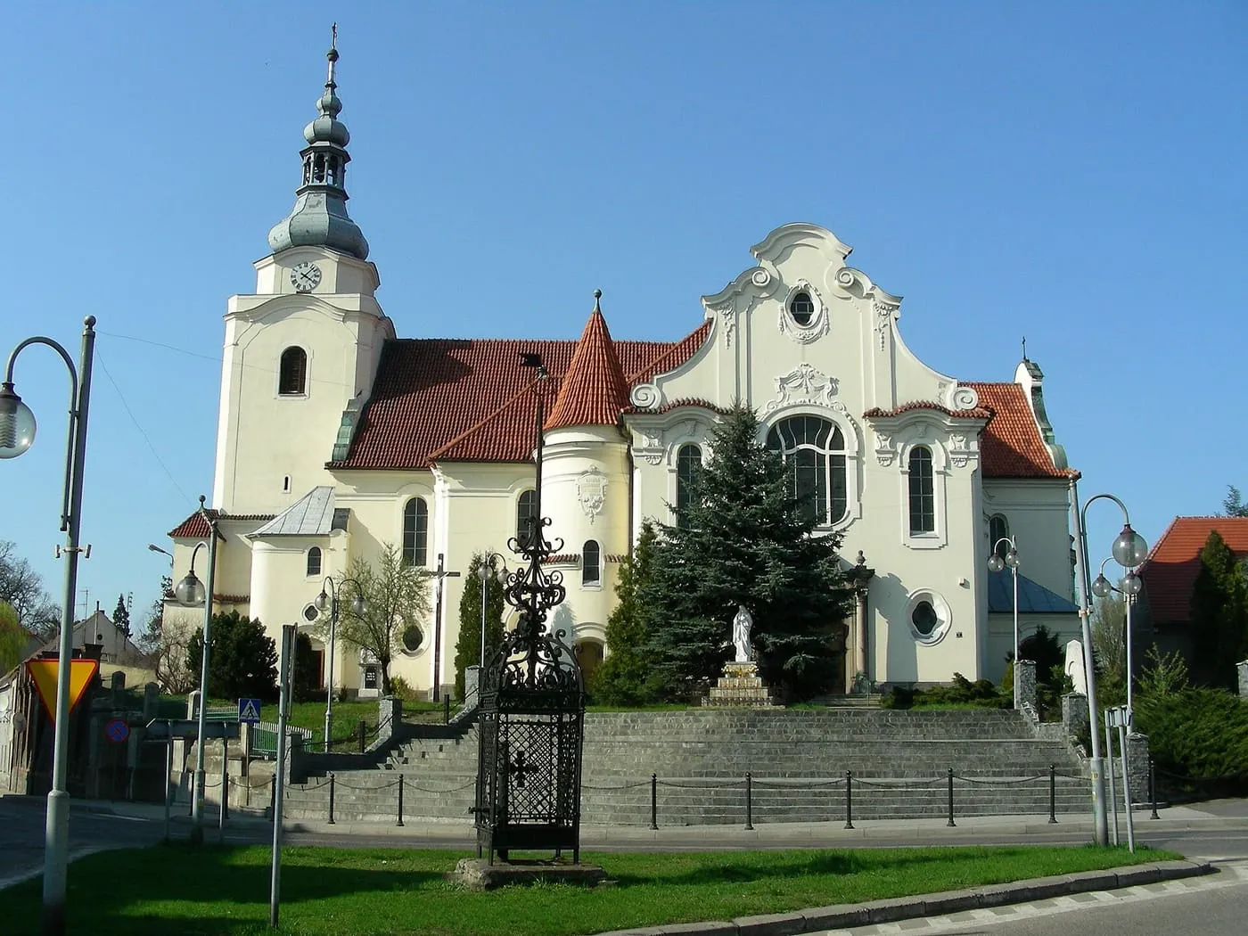 Jasny kościół z ozdobnym, falistym szczytem fasady i wieżą zwieńczoną zielonkawym hełmem, stojący na wzniesieniu za murowanym ogrodzeniem i kutą bramą, na tle błękitnego nieba