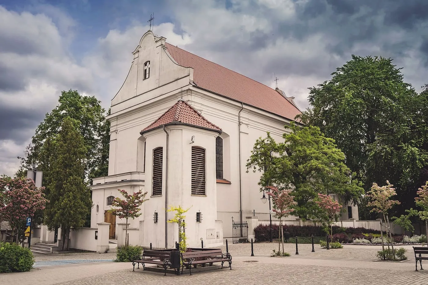 Biały, barokowy kościół z ozdobnym szczytem i czerwonym dachem, widziany z rynku spośród drzew, z ławkami na pierwszym planie i ciemnymi chmurami w tle