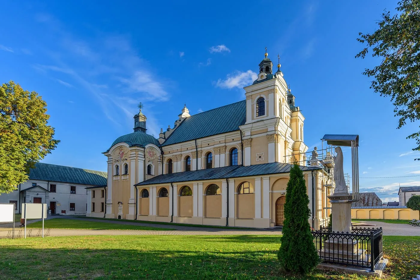 Kremowy, barokowy kościół z dwuwieżową fasadą i zielonym dachem oraz kopułą kaplicy, widziany od strony trawnika i zabudowań klasztornych pod intensywnie błękitnym niebem